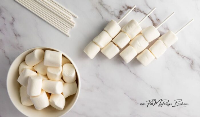 steps to make these Simple Marshmallow Pops for Valentine's idea for kids. Stack three marshmallows on a stick, dip in chocolate, and add heart sprinkles.