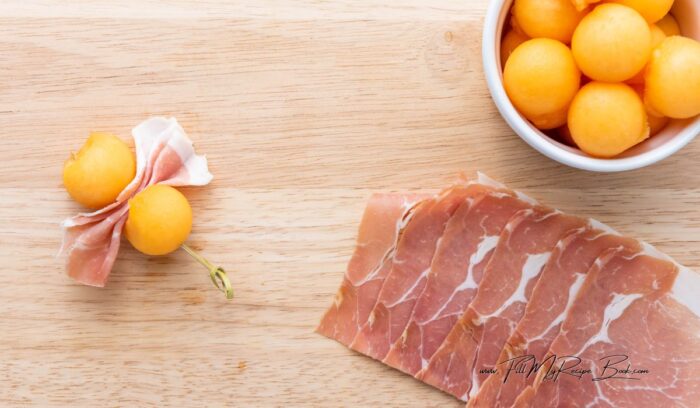 then carefully thread the folded prosciutto onto the skewer, placing it next to the cantaloupe. The prosciutto should sit nicely against the cantaloupe ball, showcasing the bow pattern.